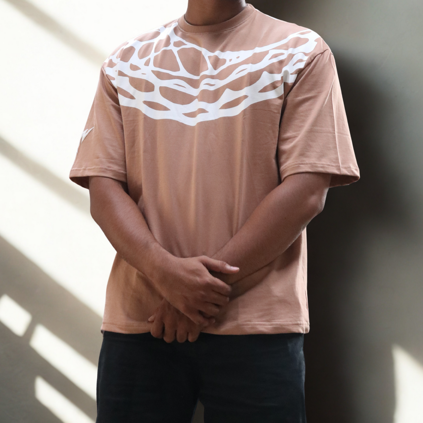 Man wearing a beige t-shirt with a white design against a wall with sunlight casting shadows.